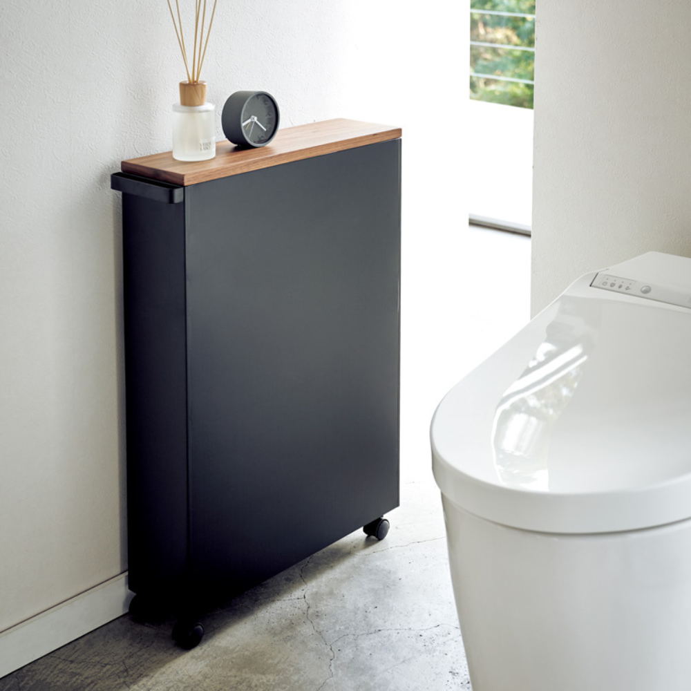 De TOWER opbergwagen met handgreep van YAMAZAKI in zwart staat met zijn houten blad en wieltjes naast een modern wit toilet in een minimalistische badkamer en erop staan een klok en een geurverspreider.