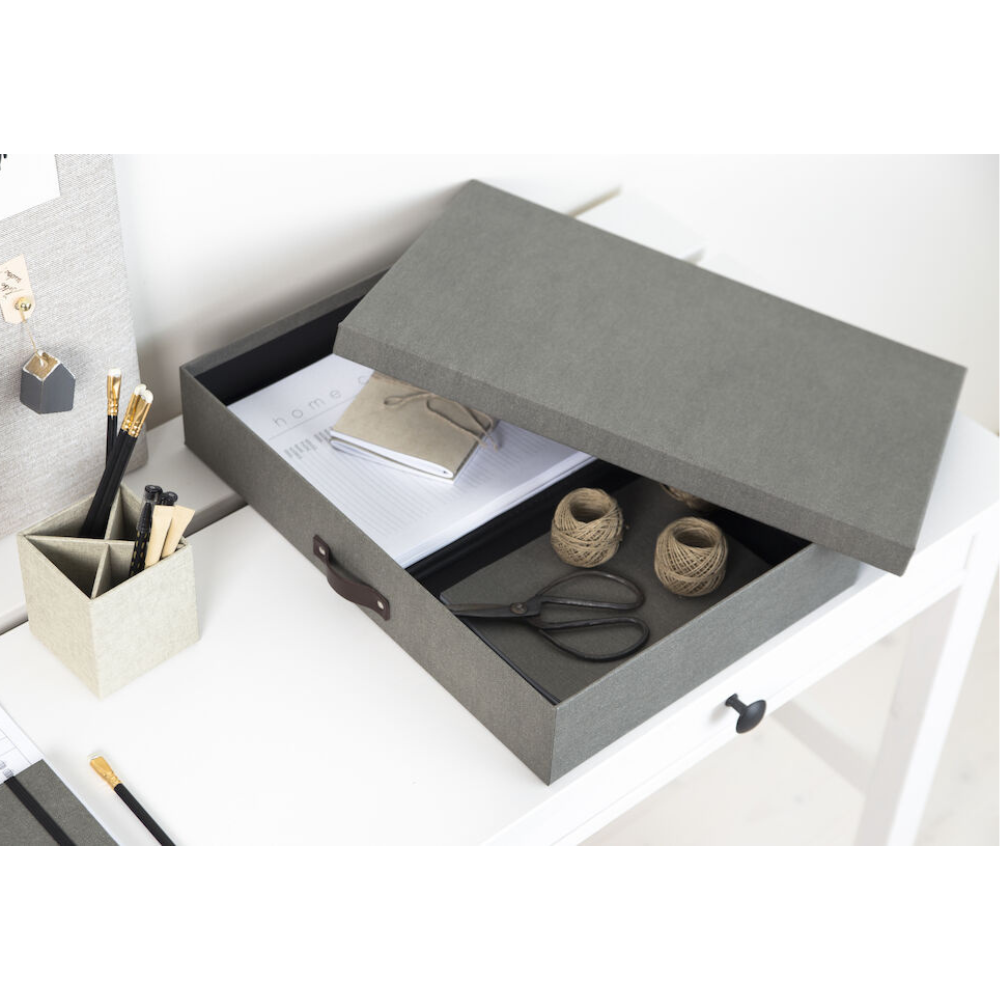 Een grijze Bigso SVERKER opbergbox A3 van canvas staat met half geopend deksel op een wit bureau. Binnenin: notitieboekjes, touw en een schaar – ideaal voor het opbergen van documenten in een georganiseerde werkplek.