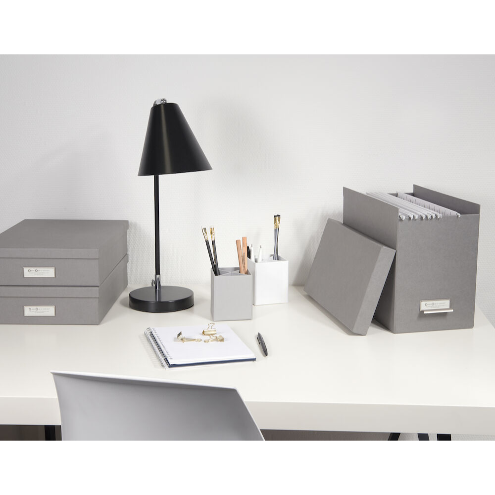 Een opgeruimd bureau met grijze opbergbakken, de Bigso JOHAN hangmappenbak – grijs voor een efficiënte documentorganisatie, een ordner, een lamp, twee pennenhouders met pennen en potloden, een open notitieboekje met pen en een witte stoel ervoor.