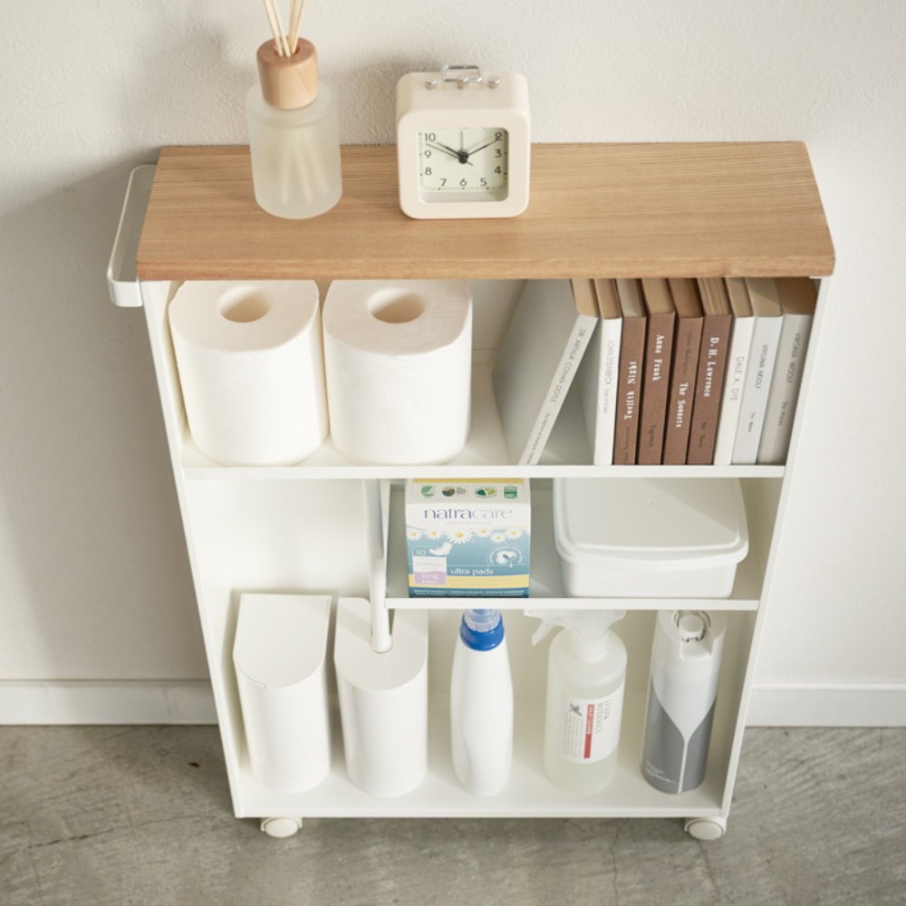 De YAMAZAKI TOWER opbergwagen met handgreep in wit, met houten blad, staat tegen een witte muur op een grijze vloer en biedt extra opbergruimte in de badkamer voor een klok, een diffuser, toiletpapier, boeken en nog veel meer.