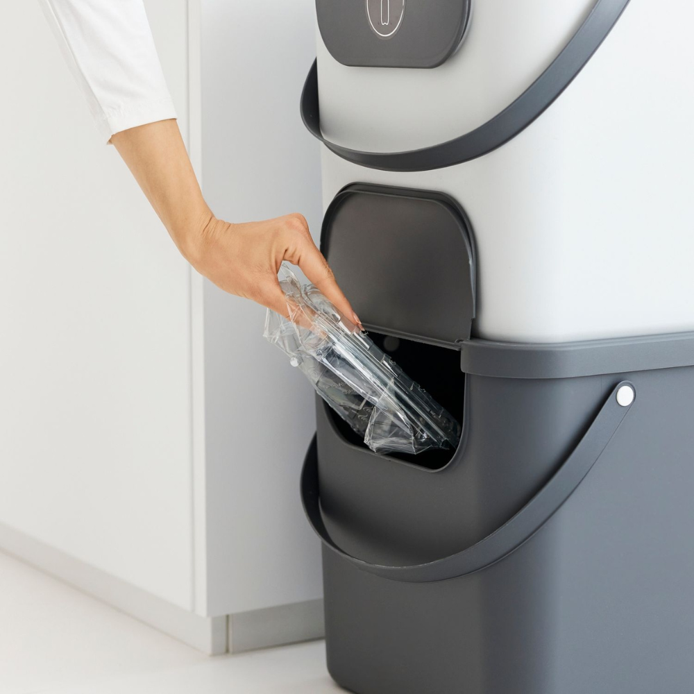 Iemand plaatst een doorzichtige plastic bak in een Rotho ALBULA-recyclingbak van 40 liter – antraciet – met een opening aan de zijkant.