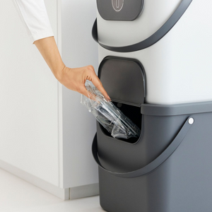 Iemand plaatst een doorzichtige plastic bak in een Rotho ALBULA-recyclingbak van 40 liter – antraciet – met een opening aan de zijkant.
