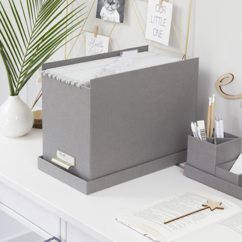 Een grijze JOHAN-hangmappenbox van Bigso met A4-hangmappen staat op een wit bureau naast een bijpassende organizer en pennen; een openliggend notitieboekje, een stervormige pen en decoraties maken deze opgeruimde werkplek compleet.