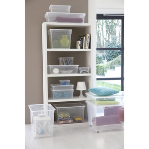 Op een witte plank staan boeken, vazen, een lamp en een Rotho-opbergdoos CLEAR 5L (33,5 x 19 x 11 cm) met allerlei spulletjes. Andere Rotho-dozen – sommige gevuld met kussens of speelgoed – staan opgestapeld op de vloer naast een groot raam.