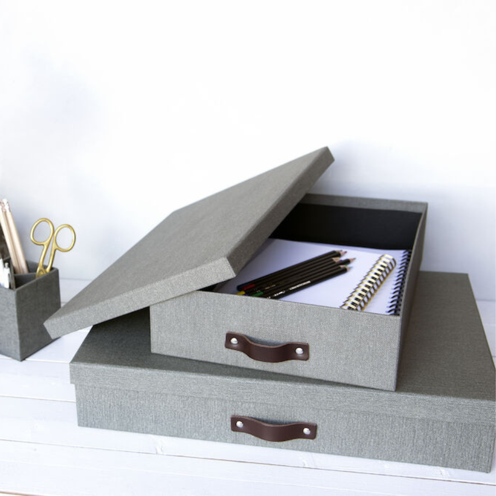 Een grijze Bigso SVERKER opbergbox A3 van canvas met een handvat ligt open op een wit oppervlak en laat zien dat er notitieboekjes en pennen in zitten. Daaronder staat nog een gesloten box met een klein bakje met pennen en een schaar ernaast.