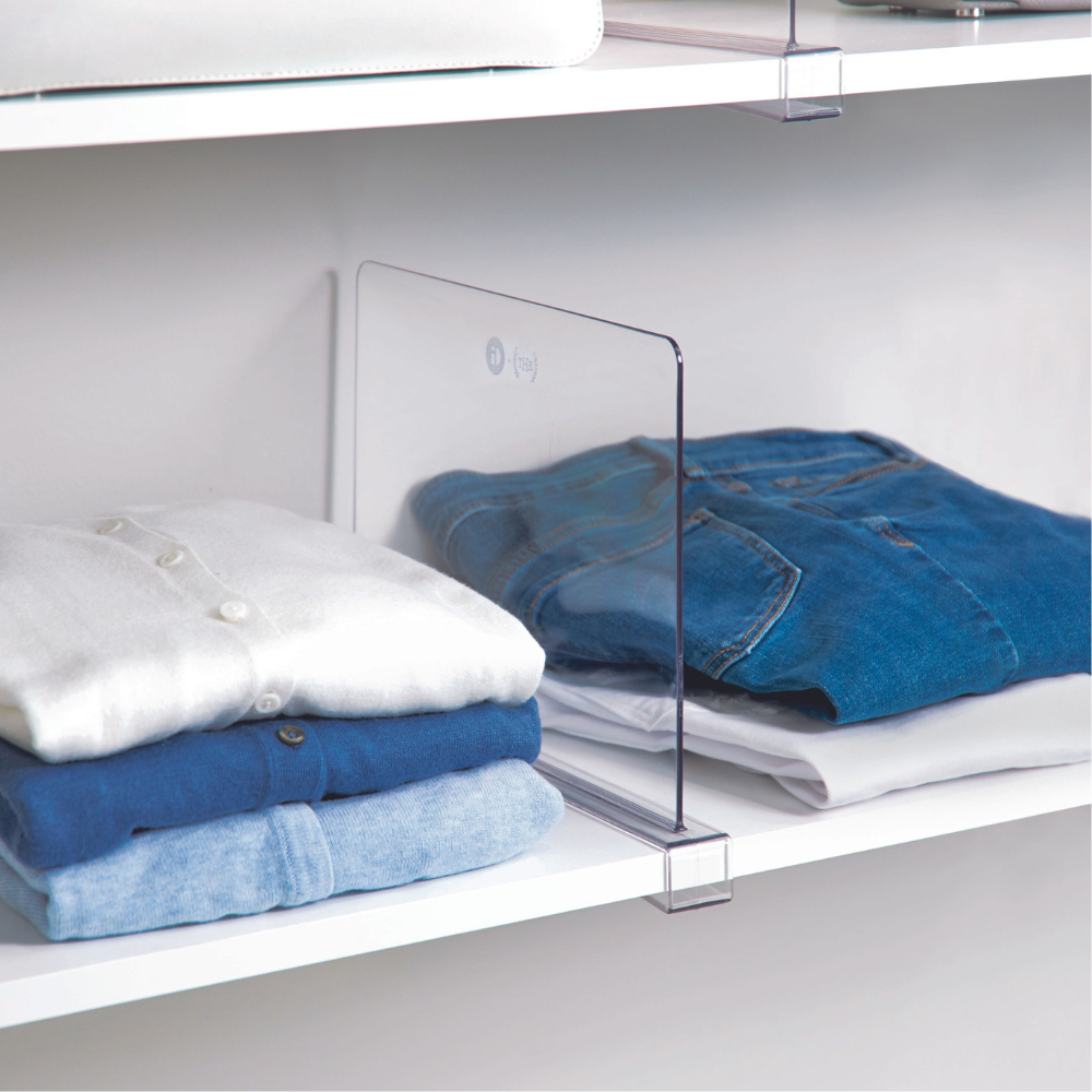 Drie netjes opgevouwen stapels witte, blauwe en spijkerkleurige kleding worden op een witte plank gescheiden door de doorzichtige plankverdeler HOME EDIT - Regaltrenner KLAR van iDesign.