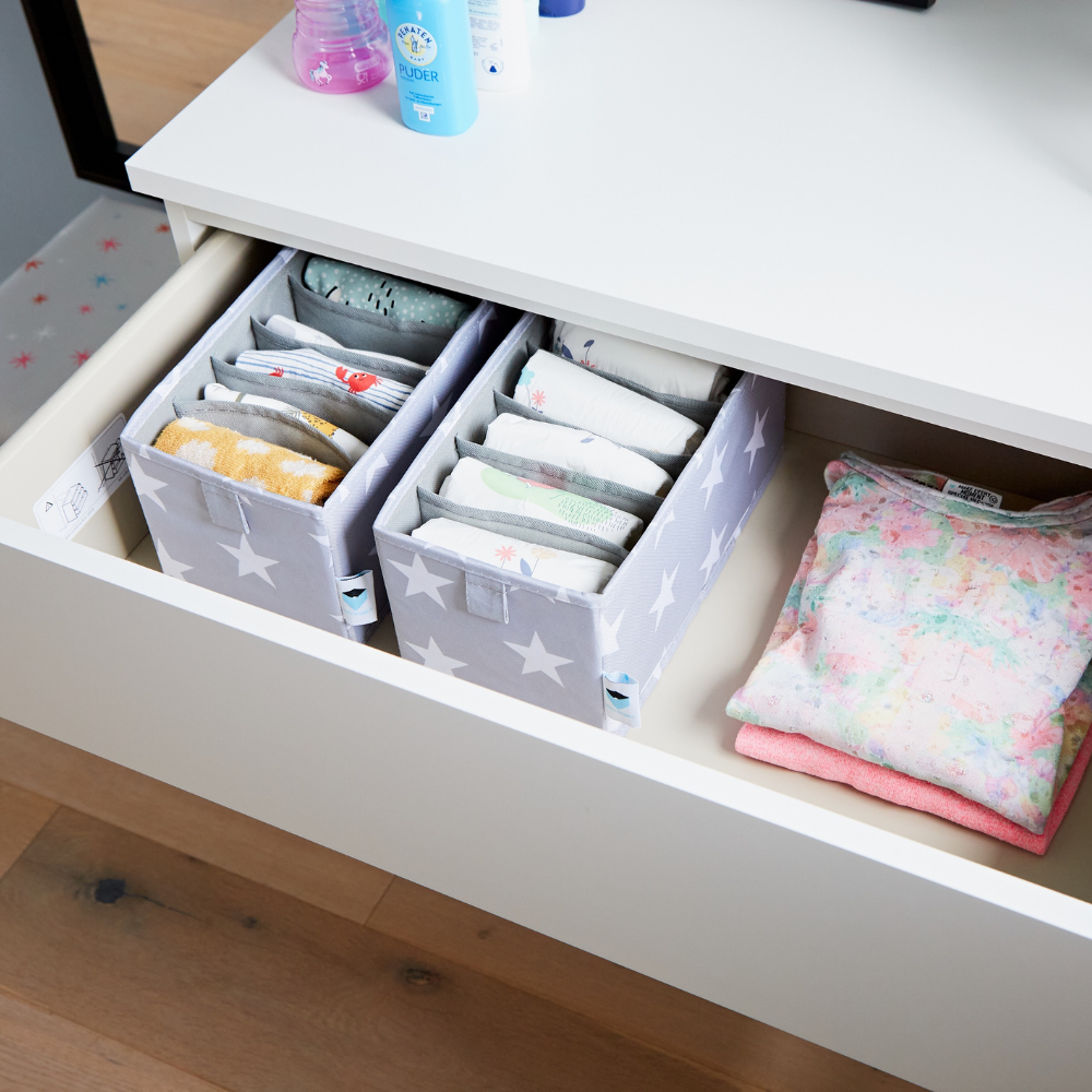 In een open witte lade liggen 2 Love it Store it STAR-organizers met 6 vakken (smal, grijs) gevuld met babykleding en accessoires, ernaast pastelkleurige babykleding en daarboven netjes gerangschikte toiletartikelen – ideaal voor de kinderkamer.