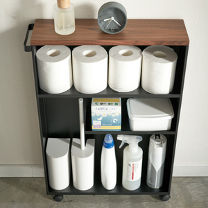 De TOWER opbergwagen met handgreep van YAMAZAKI in zwart biedt op drie planken ruimte voor vijf rollen toiletpapier, een klein klokje, flessen, een doos maandverband, een bakje en schoonmaakspullen aan een witte wand.