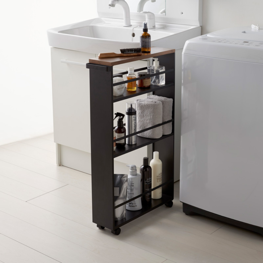 De YAMAZAKI TOWER-keukentrolley met handgreep in zwart, gevuld met toiletartikelen, handdoeken en flessen, staat tussen de witte wastafel en de wasmachine in de minimalistische badkamer en zorgt voor slimme opbergruimte.
