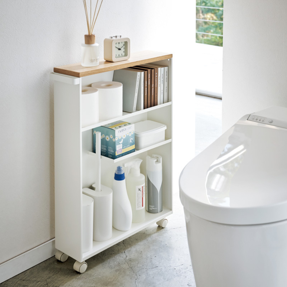 De YAMAZAKI TOWER opbergwagen met handgreep in wit, die naast een modern wit toilet staat, biedt netjes ruimte voor schoonmaakmiddelen, toiletpapier en boeken. Een klok, een geurverspreider en een dienblad bovenop zorgen voor een slimme opbergruimte in de lichte badkamer.