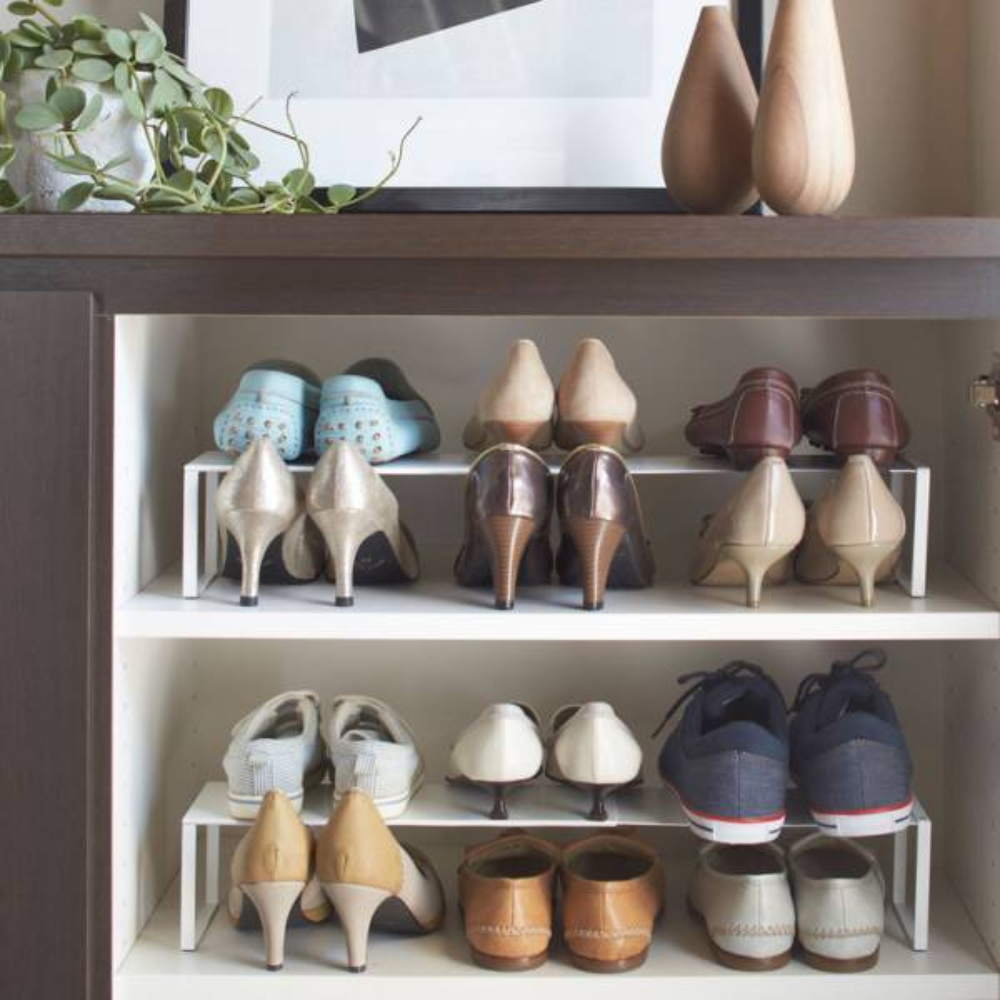 De uitschuifbare FRAME schoenenkast - Wit van YAMAZAKI biedt ruimte aan hoge hakken, platte schoenen en sneakers op witte planken onder een donkere houten kast; bovenin zorgen vazen, planten en kunst voor elegante accenten in deze slimme opbergruimte.