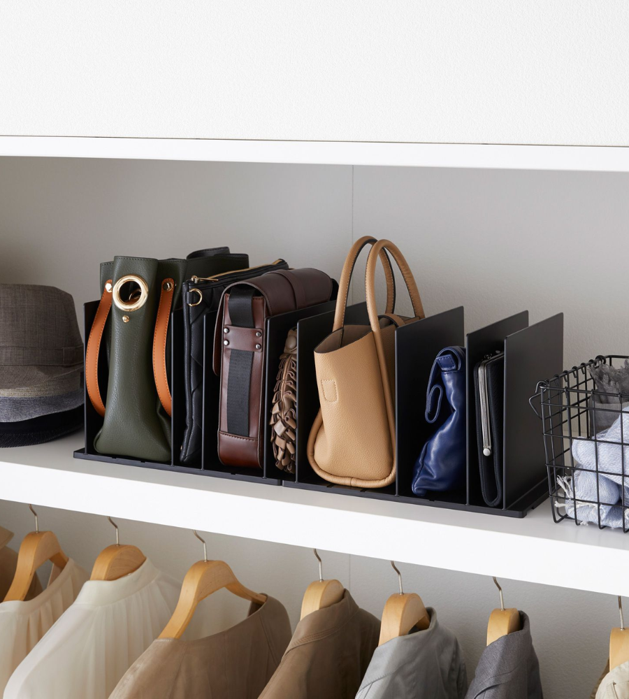 Meerdere handtassen en clutches liggen netjes opgeborgen in twee zwarte YAMAZAKI TOWER-tasorganizers (set van 2 stuks) met verstelbare scheidingswanden op een witte kastplank, naast hoeden en opgevouwen kleding. Daaronder hangen overhemden.