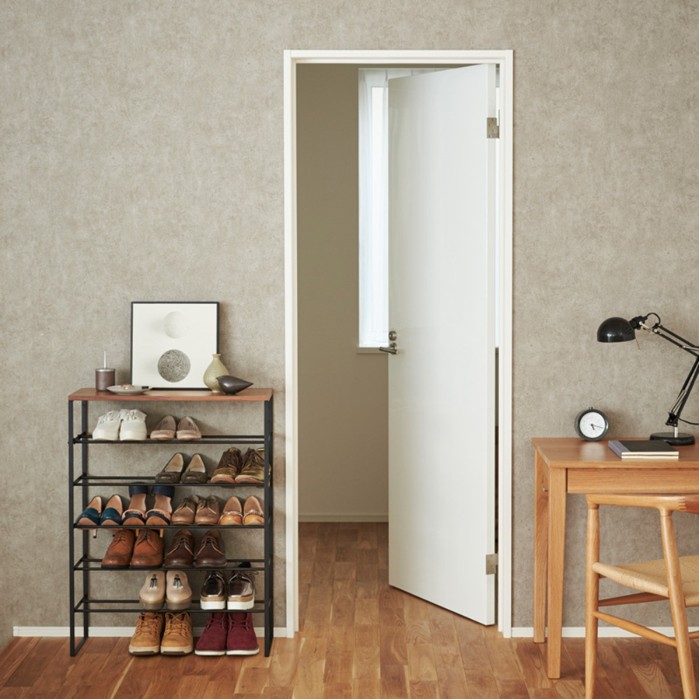 In een opgeruimde kamer staat de YAMAZAKI TOWER schoenenrek van hout - zwart, een ruimtebesparend schoenenrek met zes planken waarop schoenen, ingelijste kunst en decoratie staan. Vlakbij staat een houten bureau met een lamp, een klok, een stoel en een witte deur die uitkomt op een licht raam.