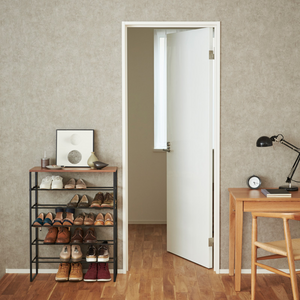 In een opgeruimde kamer staat de YAMAZAKI TOWER schoenenrek van hout - zwart, een ruimtebesparend schoenenrek met zes planken waarop schoenen, ingelijste kunst en decoratie staan. Vlakbij staat een houten bureau met een lamp, een klok, een stoel en een witte deur die uitkomt op een licht raam.