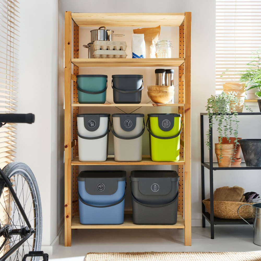 Op een houten plank staan kleurrijke bakjes, keukengerei en de Rotho 3-delige set ALBULA Recycling-afvalsysteem 25 l in antraciet. Met planten en een fiets in de buurt zorgt het zonlicht dat door de jaloezieën schijnt voor een gezellige ruimte die het scheiden van afval stimuleert.