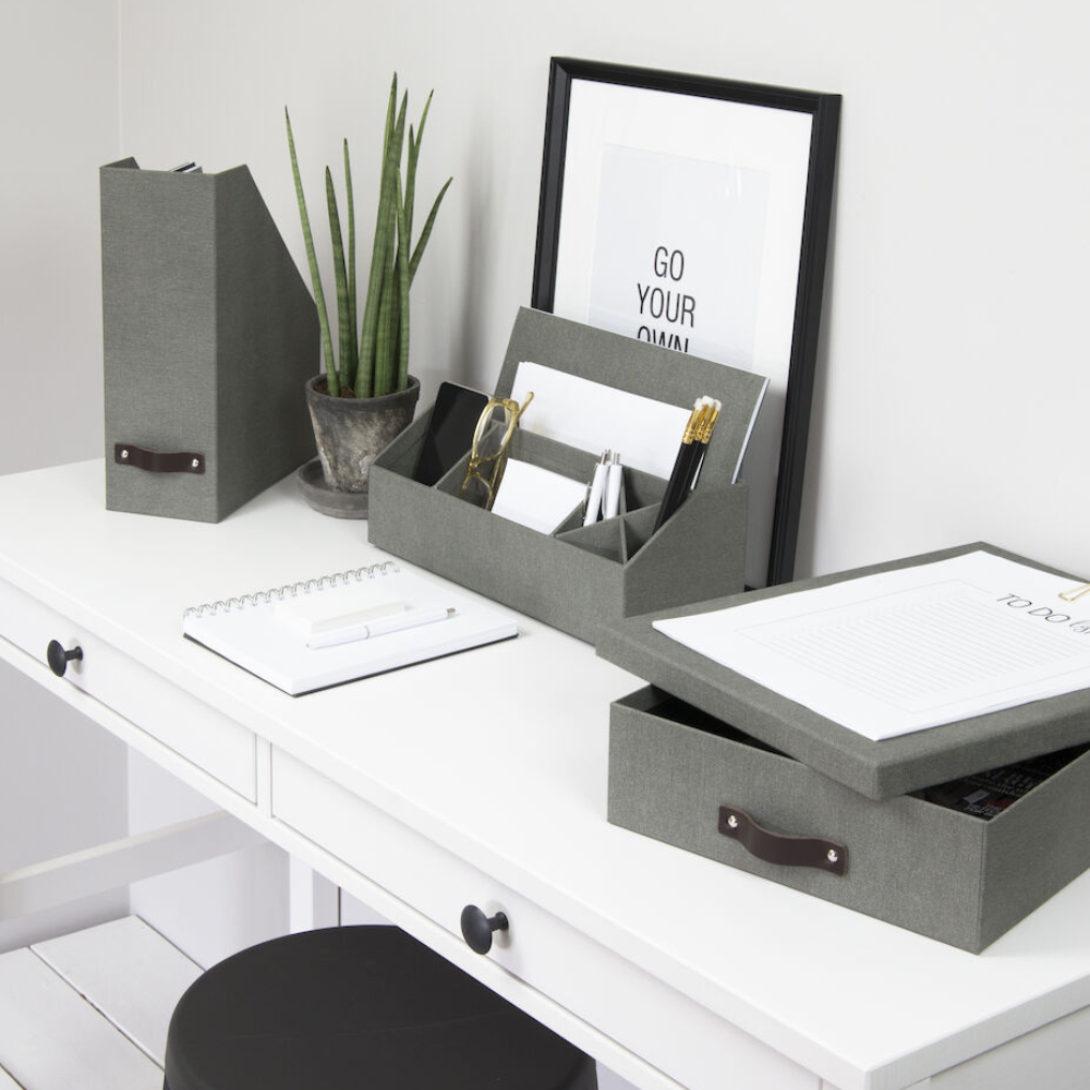 Een opgeruimd wit bureau met grijze opbergers, een Bigso OSKAR opbergdoos A4 in grijs canvas, een notitieboekje, een ingelijste quote met "GO YOUR OWN WAY", een potplant en schrijfspullen die netjes zijn gerangschikt. Daaronder staat een zwarte kruk.