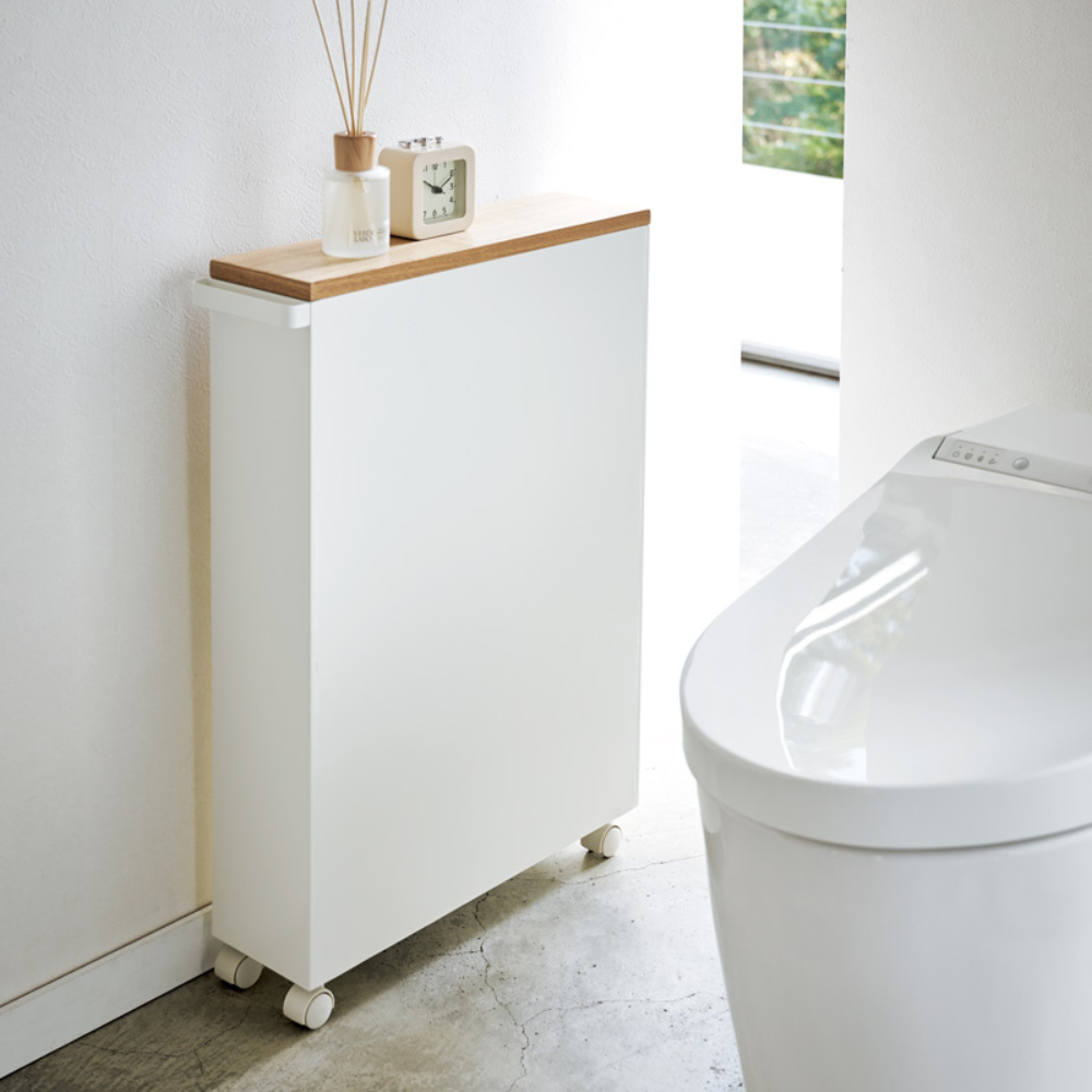 De YAMAZAKI TOWER opbergwagen met handgreep in wit, met houten plank en wieltjes, staat naast een modern wit toilet en biedt stijlvolle opbergruimte in de badkamer. Bovenop staan een klein klokje, een kamerparfum en een flesje in het zonlicht.