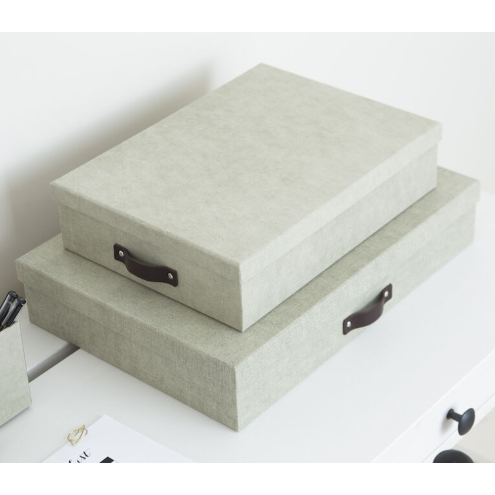 Twee op elkaar gestapelde lichtgrijze Bigso OSKAR opbergboxen A4 – linnen canvas met zwarte handvatten staan op een wit oppervlak, ideaal voor het opbergen van documenten in je thuiskantoor. Ernaast liggen een klein dienblad en wat papierwaren.