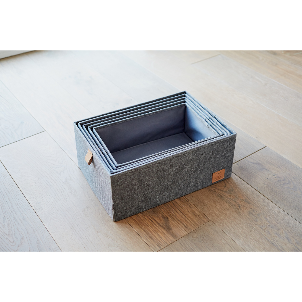 Vijf rechthoekige, grijze opbergboxen van stof uit de PREMIUM-set met 5 opbergboxen met deksel in grijs van Love it Store it staan in elkaar geschoven op een houten vloer. De grootste box heeft een klein bruin leren label – perfect om thuis meer orde te scheppen.