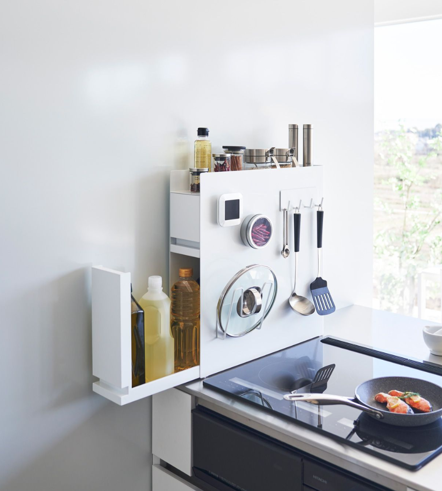 Het YAMAZAKI TOWER kruidenrek met twee verdiepingen – wit – past perfect in een moderne, minimalistische keukenhoek en houdt keukengerei, kruiden, flessen en een pannendeksel netjes geordend boven een elegante inductiekookplaat, waar het zonlicht doorheen schijnt.