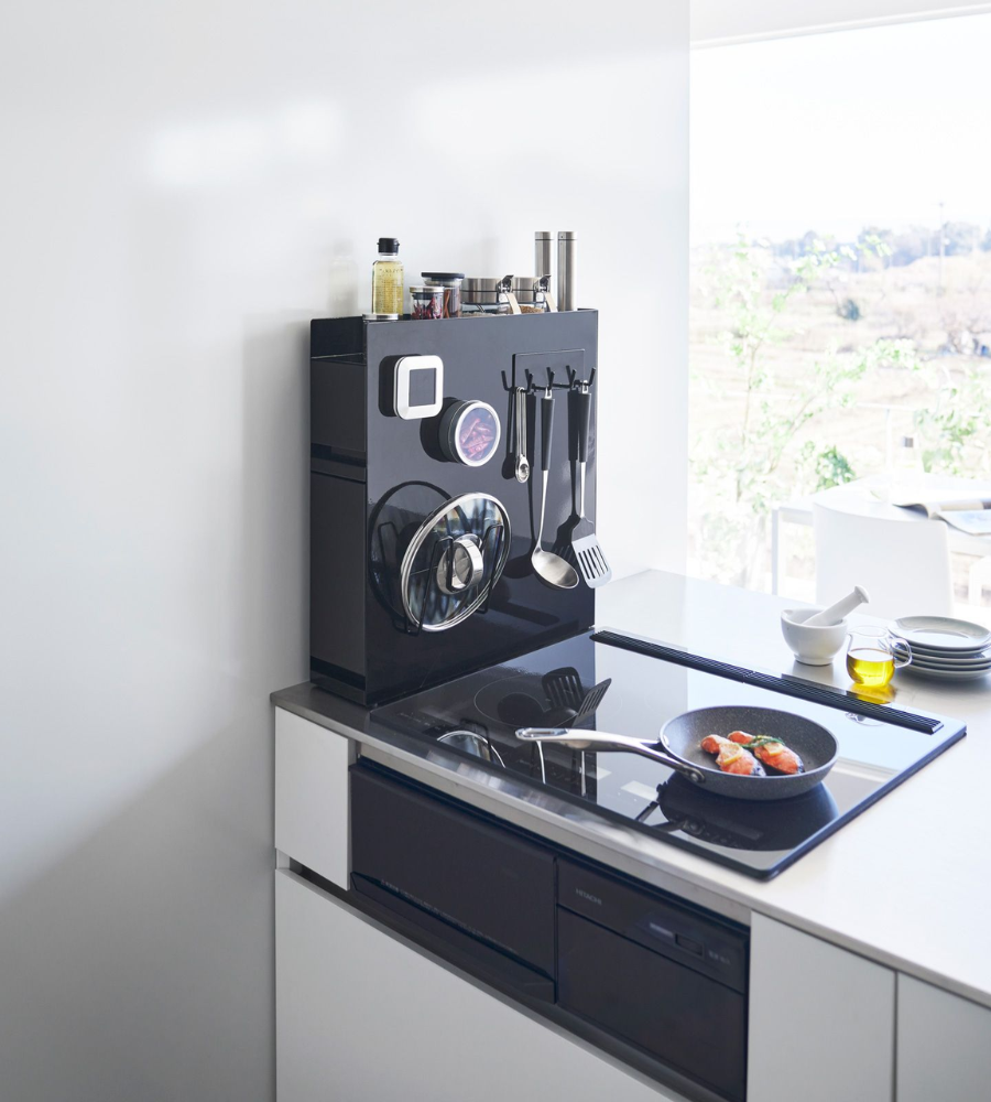 Een moderne keuken met een elegant zwart fornuis en een koekenpan toont het YAMAZAKI TOWER kruidenrek met twee verdiepingen in zwart en keukengerei op een magnetisch oppervlak. Door het raam zie je bomen in de zon.