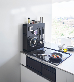Een moderne keuken met een elegant zwart fornuis en een koekenpan toont het YAMAZAKI TOWER kruidenrek met twee verdiepingen in zwart en keukengerei op een magnetisch oppervlak. Door het raam zie je bomen in de zon.