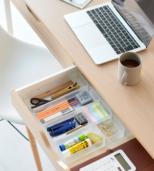 In een open lade, uitgerust met de YAMAZAKI TOWER lade-organizer wit L (set van 2 stuks), is er ruimte voor pennen, een schaar, plakbriefjes, lijmstiften, paperclips en een liniaal. Op het houten bureau erboven staan een laptop, een koffiebeker en een rekenmachine.