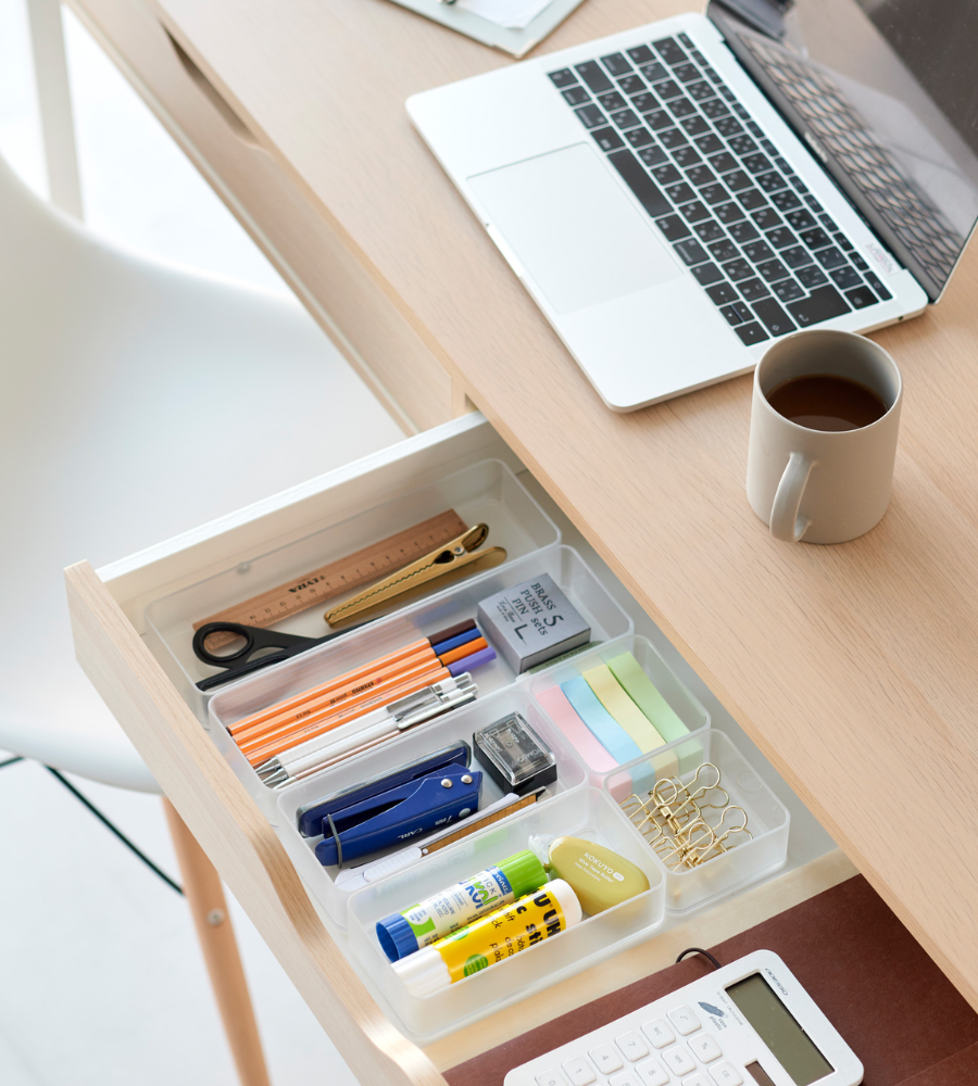 In een open lade, uitgerust met de YAMAZAKI TOWER lade-organizer wit L (set van 2 stuks), is er ruimte voor pennen, een schaar, plakbriefjes, lijmstiften, paperclips en een liniaal. Op het houten bureau erboven staan een laptop, een koffiebeker en een rekenmachine.