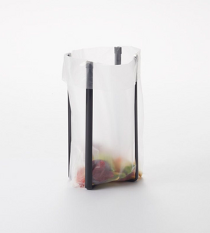 Een doorzichtige plastic zak, die open wordt gehouden door de YAMAZAKI TOWER multifunctionele keukenstandaard – zwart, een elegante zwarte metalen standaard, laat de kleurrijke spullen binnenin zien tegen een eenvoudige witte achtergrond.