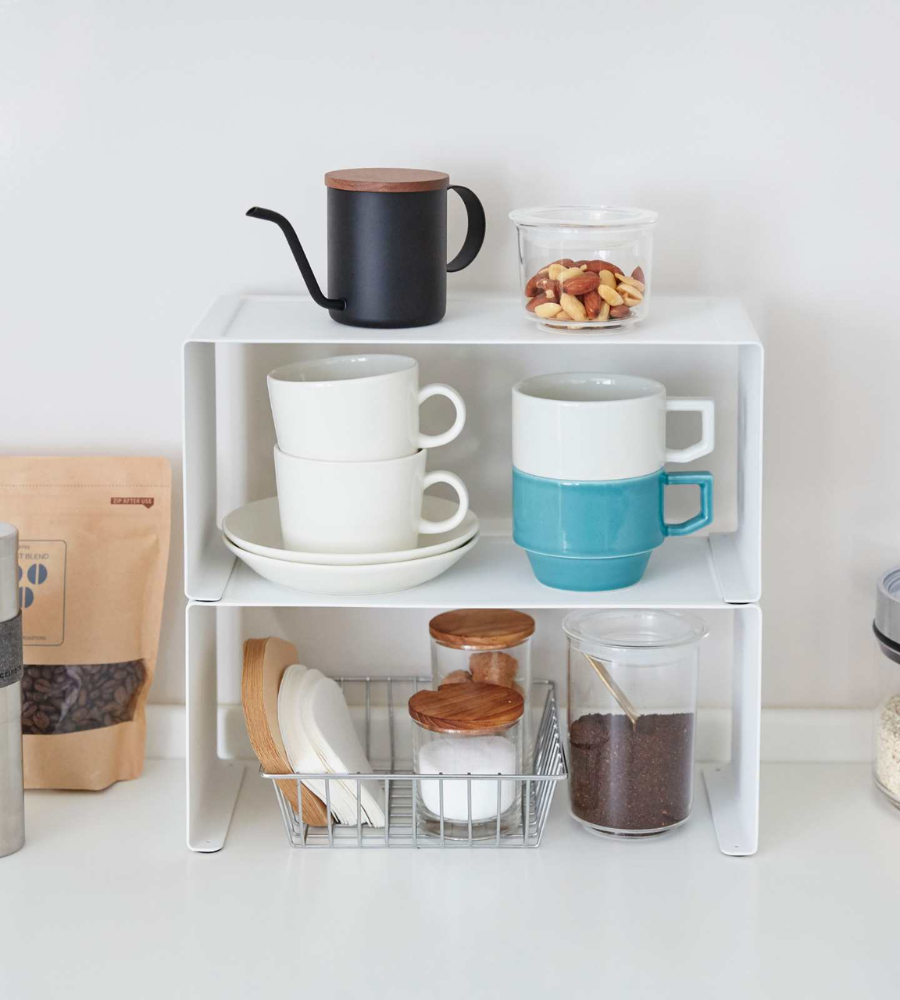 De smalle TOWER-keukenkast van YAMAZAKI in wit biedt ruimte aan stapelbare kopjes, een zwarte waterkoker, een pot met noten, een draadmand met borden, twee glazen met houten deksels en koffiepotjes – een perfect gebruik van de ruimte in de keuken.