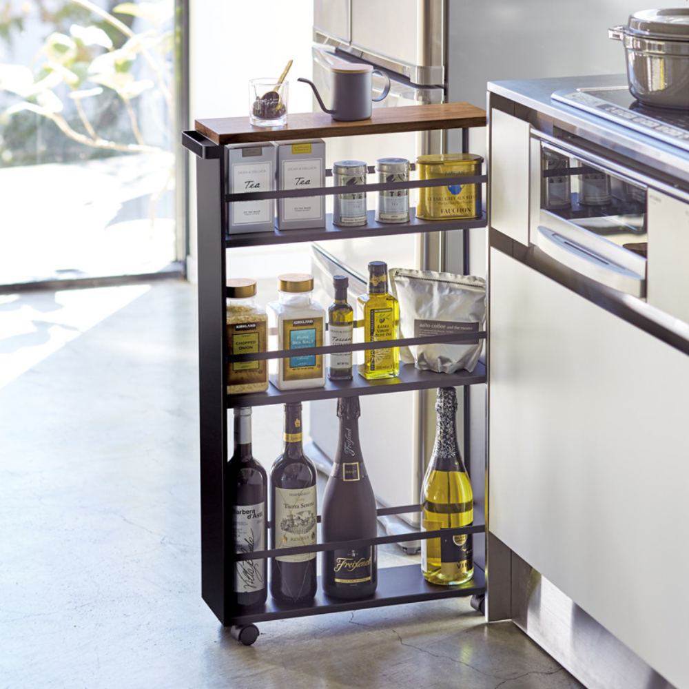 De YAMAZAKI TOWER keukentrolley met handgreep in zwart is een slanke metalen trolley met vier schappen, ideaal voor olie- en azijnflessen, kruidenpotjes en nog veel meer naast het moderne keukenkastje bij het raam.