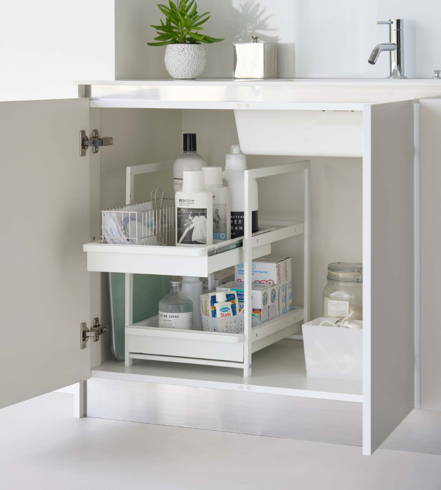 In de open badkamerkast onder de wastafel staan flessen, lotions en schoonmaakmiddelen netjes opgeruimd op een YAMAZAKI TOWER laderek met 2 verdiepingen – wit. Een klein plantje siert het aanrecht erboven.