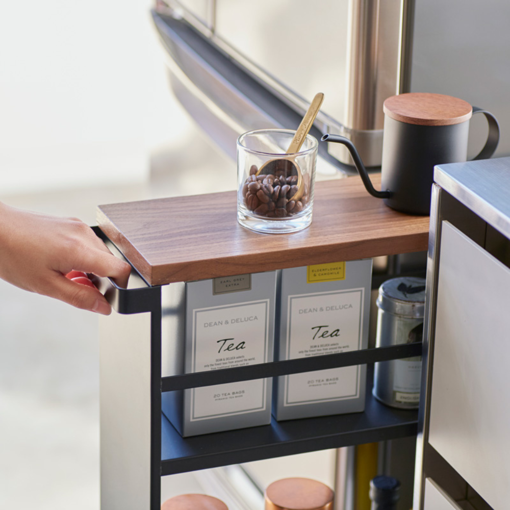 Een hand trekt het YAMAZAKI TOWER keukentrolleytje met een zwarte handgreep, waarop op het houten blad koffiebonen, een lepel en een zwarte theepot staan. Theeblikken op het onderste schap zorgen voor handige opbergruimte.