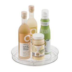 Op de iDesign HOME EDIT - draaiplateau Lazy Susan TRANSPARANT (23x3,5 cm) staan twee flessen azijn, een bus avocadospray en een potje geklaarde boter tegen een eenvoudige witte achtergrond.