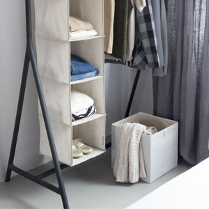 Een hangende opbergzak van stof met inlegvellen biedt ruimte voor kleding en schoenen. Ernaast staat een Bigso SOFT opbergbox CUBE in beige, met daarin een opgevouwen trui, terwijl er naast een aantal overhemden en jassen aan een zwarte kledingstandaard hangen.