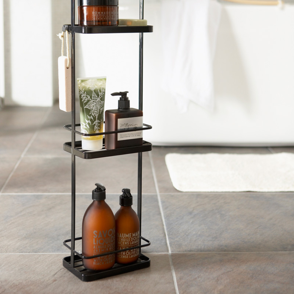 De YAMAZAKI TOWER badkamerplank in zwart biedt slimme opbergruimte voor zeepdispensers, een tandenborstelbeker, een spons en badkamerbenodigdheden op de tegelvloer, naast een witte badmat en het bad.
