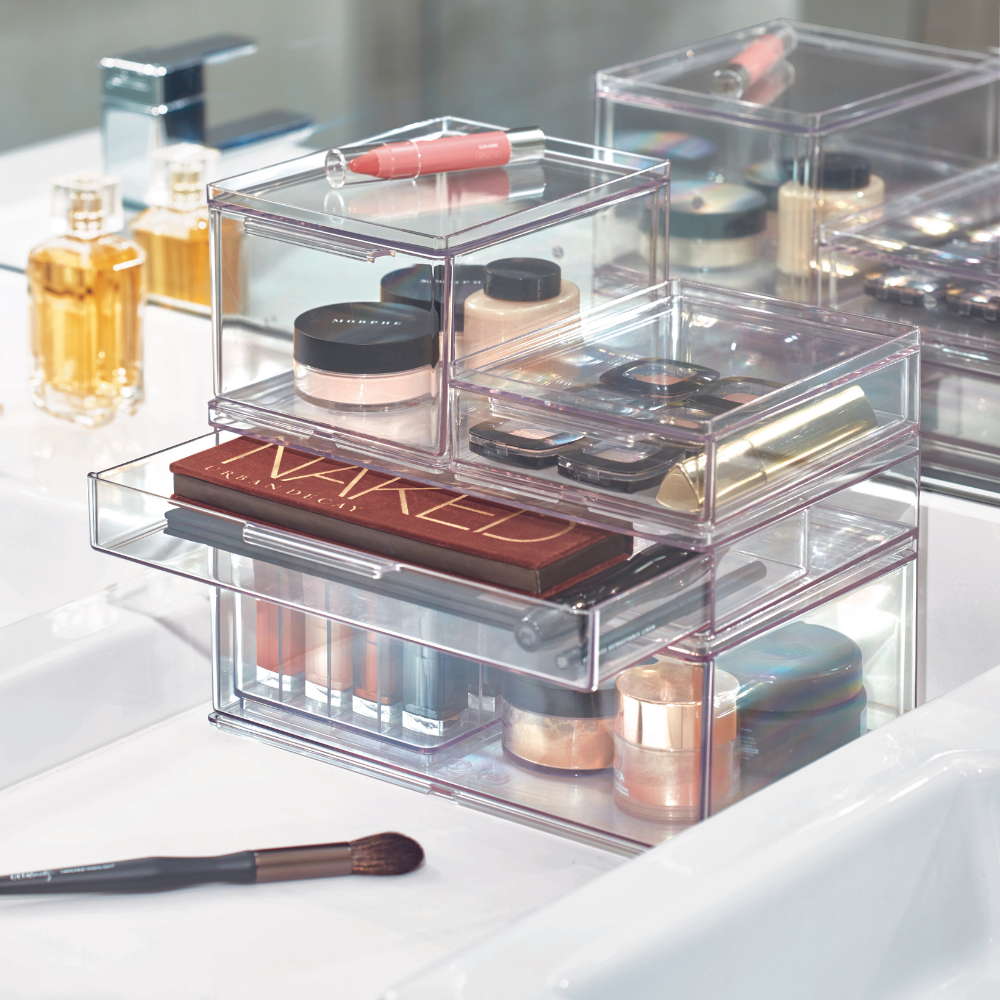 Een modulaire HOME EDIT-organizer met lade van iDesign, in verschillende maten, gevuld met paletten, foundation, lippenstift en kwasten, staat naast parfumflesjes en een make-upkwast op het witte badkamerblad.