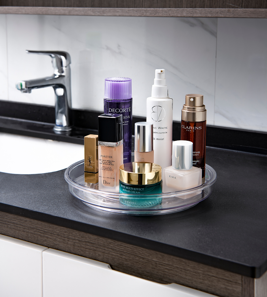 De draaiplateau Lazy Susan KLAR (23,5 x 3,7 cm) van BNB x VCN staat op een zwart badkamertafeltje en houdt huidverzorgings- en cosmeticapotjes netjes op een rijtje naast een wastafel met een zilveren kraan en een marmeren rand.