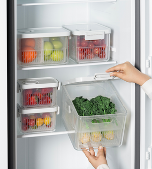 Iemand bewaart groene bladgroenten in een doorzichtige DATEMATE-koelbox KLAR Wit met houdbaarheids-timer van BNB x VCN in de koelkast. Dankzij het stapelbare ontwerp zorgen meerdere van deze boxen ervoor dat fruit en groenten overzichtelijk op de plank liggen.