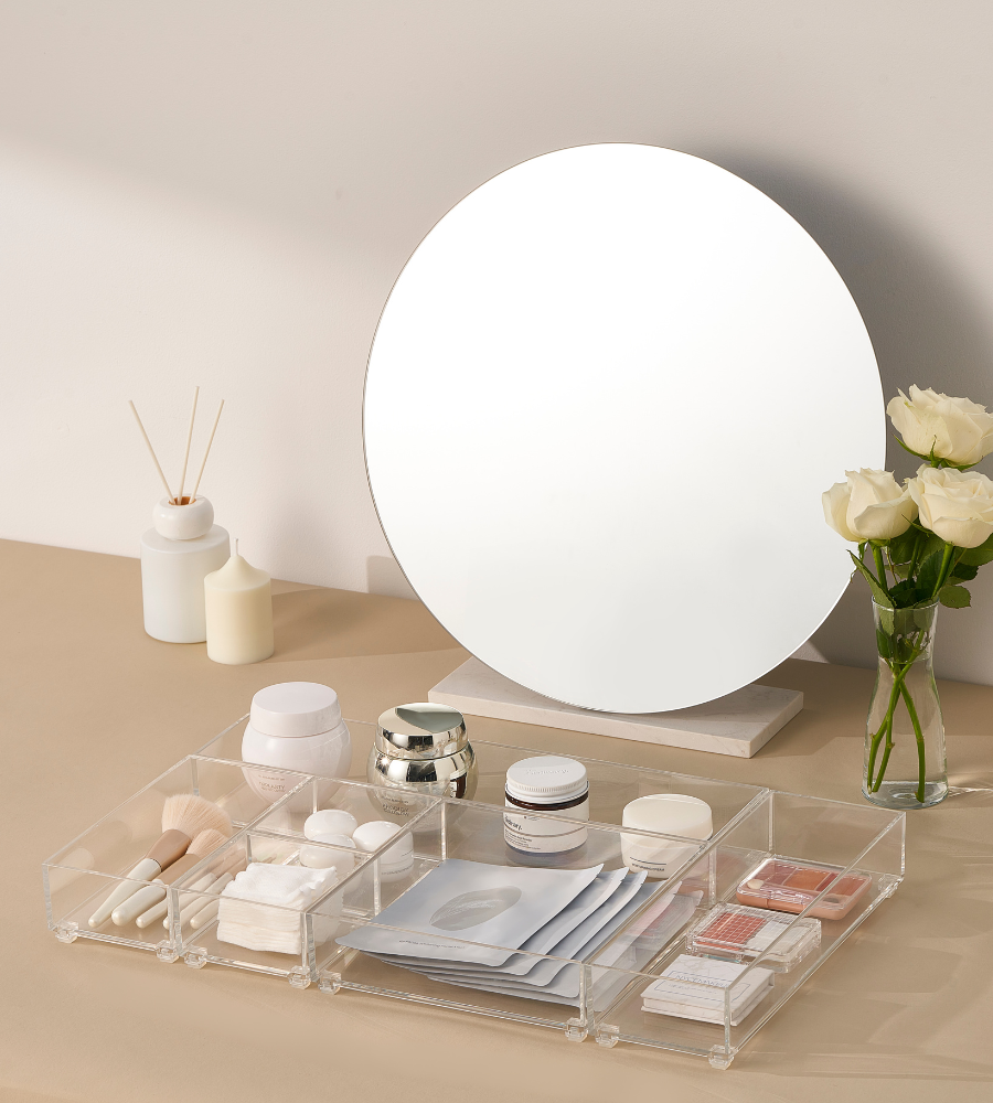 Op een beige wastafel met witte rozen en kaarsen staat een ronde spiegel. Make-upborstels, crèmes, verzorgingsproducten en gezichtsmaskers zijn netjes opgeborgen in een organizer en de BNB x VCN lade-organizerset SLIDE (5-delig).