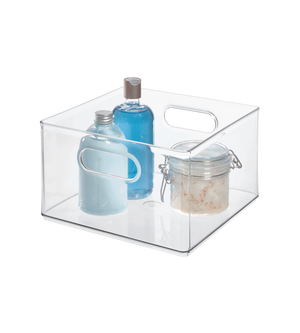 Een heldere, vierkante iDesign HOME EDIT opbergbak KLAR (25,4 x 25,4 x 15,2 cm) met handgreepuitsparingen bevat twee blauwe flessen en een pot met schuifdeksel, allemaal op een witte achtergrond.