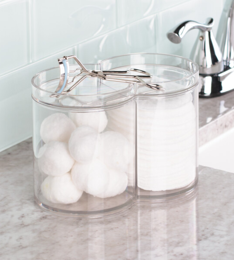 De CLARITY-cosmetica-organizer KLAR van iDesign staat, gevuld met wattenbolletjes en wattenschijfjes, op het badkamerblad. Op het deksel ligt een wimperkruller; op de achtergrond zie je de kraan en de betegelde achterwand.