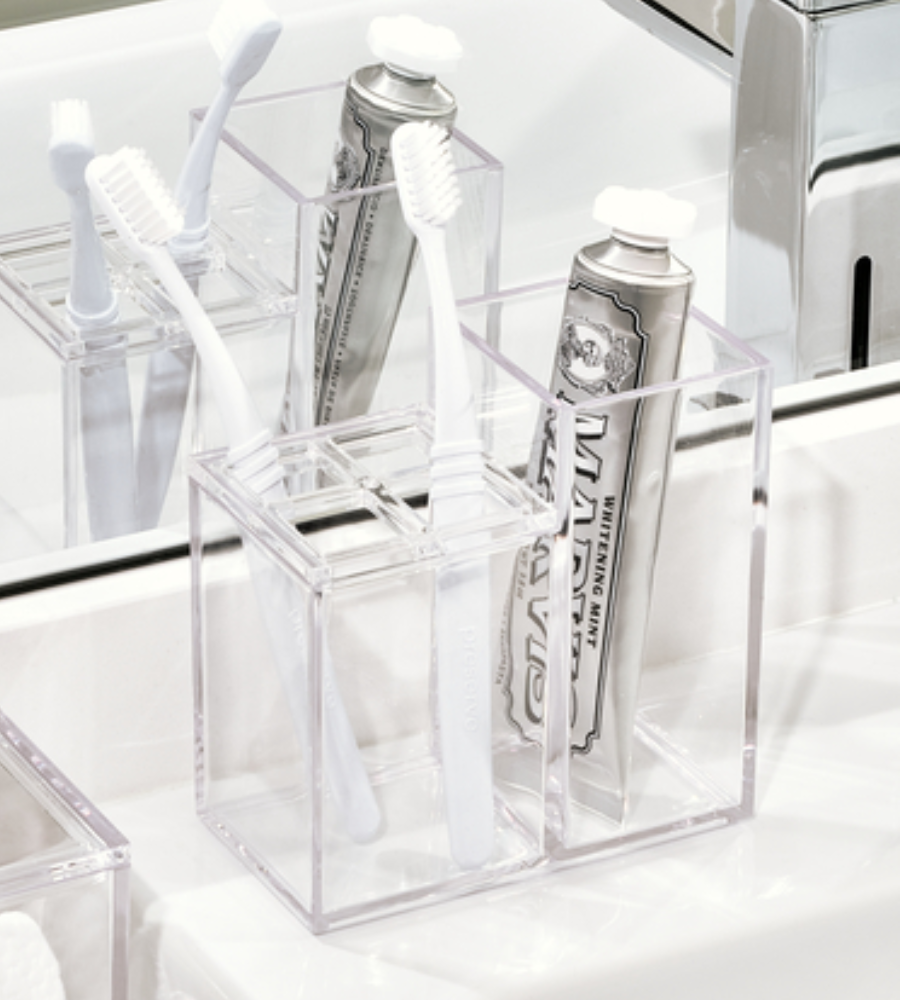 De iDesign CLARITY - Kosmetik Organizer KLAR II staat op een wastafel in de badkamer en dient als tandenborstelhouder voor twee witte tandenborstels en een zilveren tube tandpasta, waarvan de weerspiegeling zichtbaar is in de spiegel erachter.