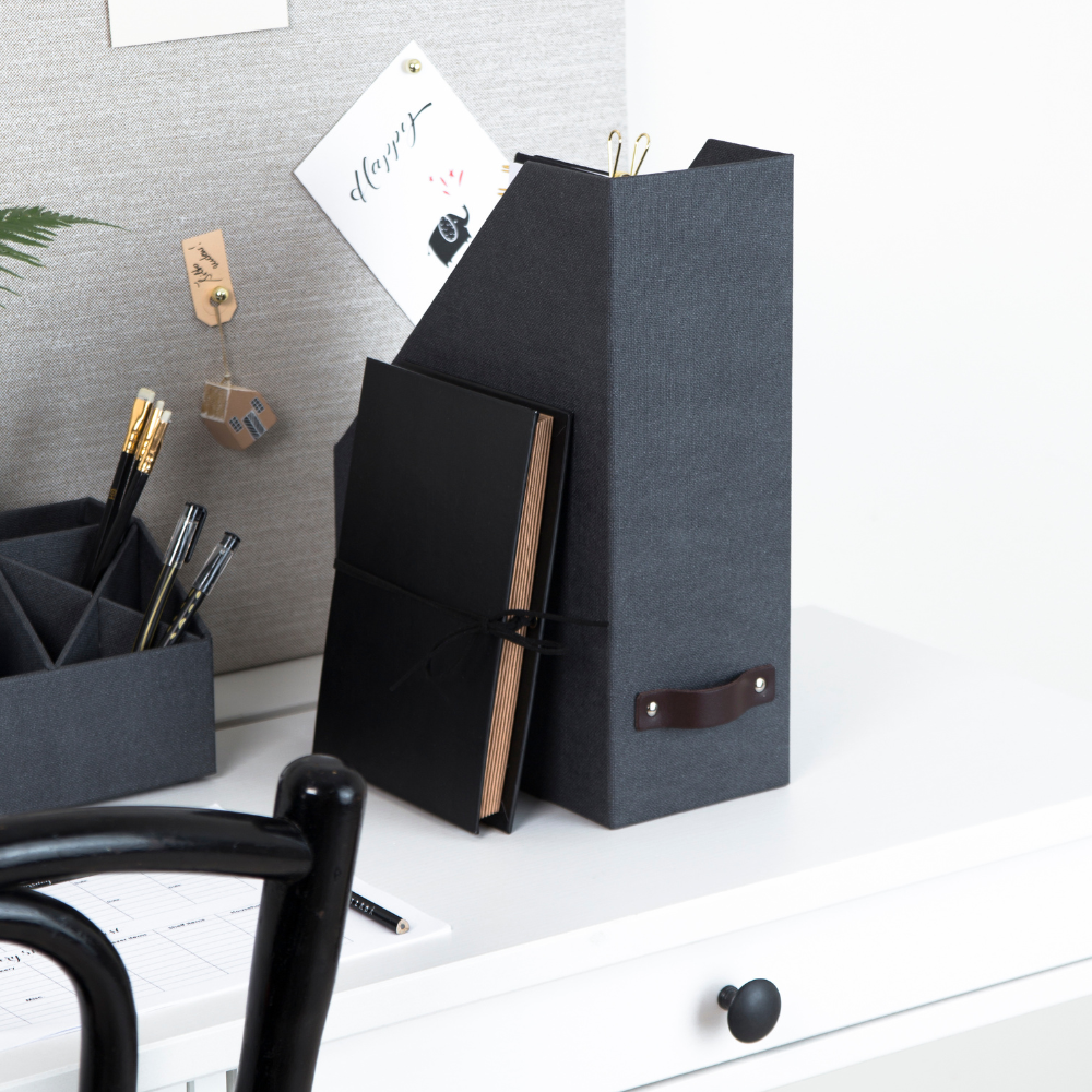 Een Bigso ESTELLE tijdschriftenhouder in zwart canvas en een notitieboekje liggen op een wit bureau naast een pennenbakje met zwarte pennen. Op de achtergrond hangt een kurkbord met notities, en op de voorgrond is een deel van een zwarte stoel te zien.