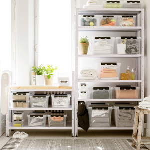 Op open planken staat de Orthex SmartStore Box KLAR 40x30x11 cm (7,5 l) – plat, BPA-vrij en voorzien van een label – naast opgevouwen handdoeken, toiletartikelen en potplanten in een lichte kamer met een tapijt met patroon.