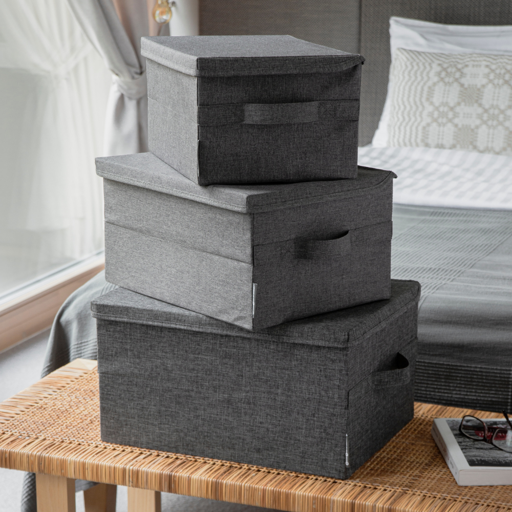 Drie Bigso SOFT opbergboxen Grijs M (40x30x22 cm) staan in de slaapkamer op een gevlochten bank gestapeld. Op de achtergrond zie je een net opgemaakt bed en een groot raam.