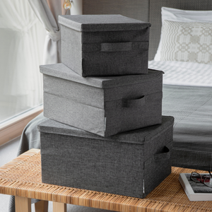 Drie Bigso SOFT opbergboxen Grijs M (40x30x22 cm) staan in de slaapkamer op een gevlochten bank gestapeld. Op de achtergrond zie je een net opgemaakt bed en een groot raam.