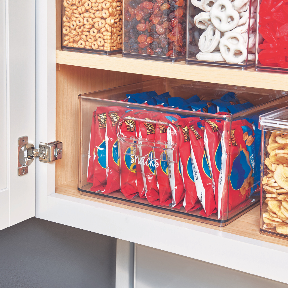 Een doorzichtige iDesign HOME EDIT-etikethouder (KLAR) met het opschrift "Snacks" bevat rode snackzakjes in een voorraadkast, omringd door andere beschrijfbare bakjes met muesli, gedroogd fruit, pretzels en chips.