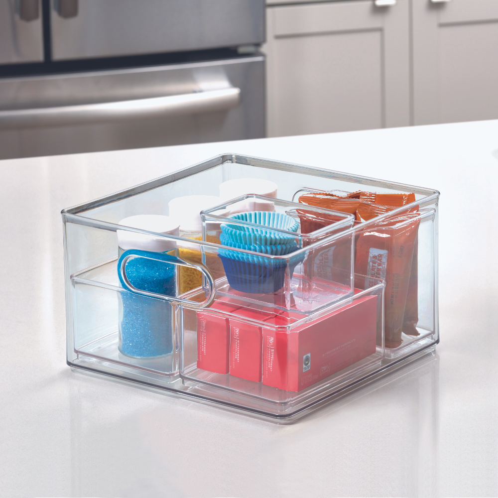 De iDesign HOME EDIT opbergbak TRANSPARANT (25,4 x 25,4 x 15,2 cm) staat op het aanrecht en houdt blauwe muffinvormpjes, kleurrijke hagelslag, rode doosjes en oranje pakjes netjes gesorteerd in aparte, stapelbare vakjes.