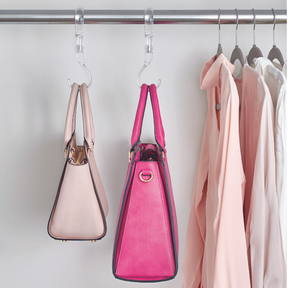 Twee handtassen, een lichtroze en een felroze, hangen ruimtebesparend aan iDesign HOME EDIT - S-haken KLAR aan een metalen stang naast een aantal lichte overhemden op kleerhangers voor een witte muur.